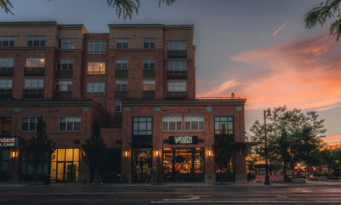 A modern brick building at sunset, featuring a restaurant with outdoor seating and trees lining the street.
