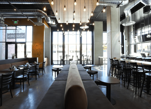 Modern restaurant interior with industrial decor, large windows, and a mix of tables and booths. Warm lighting hangs above.