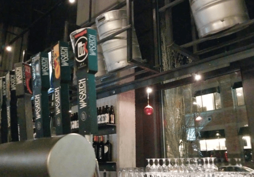 A bar scene featuring beer taps, a hanging ornament, and kegs above, with a view of city lights through the window.