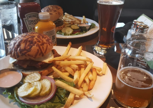 A plate with a burger, fries, pickles, and sauces, alongside a glass of beer in a casual dining setting.
