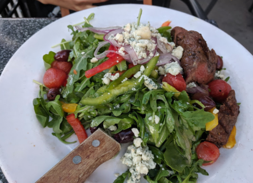 A vibrant salad with mixed greens, red onions, peppers, grapes, and blue cheese, served with a piece of grilled meat.