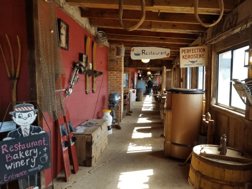 A rustic hallway featuring signs for a restaurant, bakery, and winery, with wooden decor and vintage tools on display.