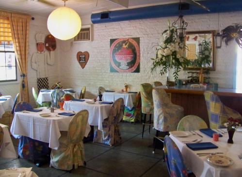 Cozy restaurant interior with colorful chairs, white tablecloths, and vibrant decor, featuring plants and artwork on the walls.