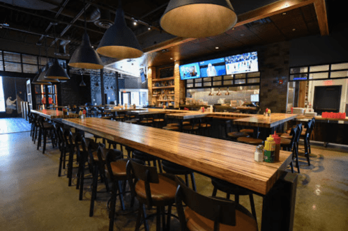 A spacious bar area with wooden tables, high chairs, and large pendant lights, featuring TVs and a modern design.