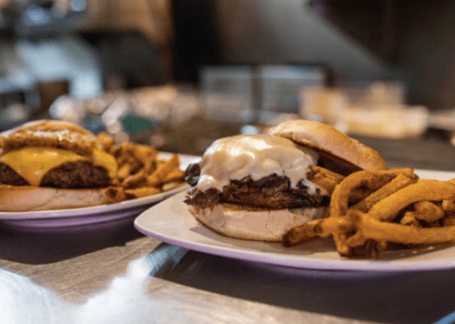 Two gourmet burgers on plates, one with cheese, accompanied by crispy fries in a restaurant setting.
