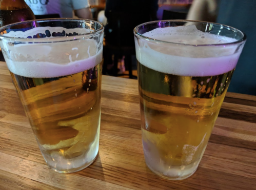 Two glasses of beer with foam on top, placed on a wooden bar surface. Colorful lights in the background.