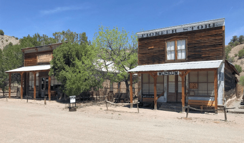 Two rustic wooden buildings labeled "Pioneer Store" and "Museum," set in a dusty outdoor area with trees.