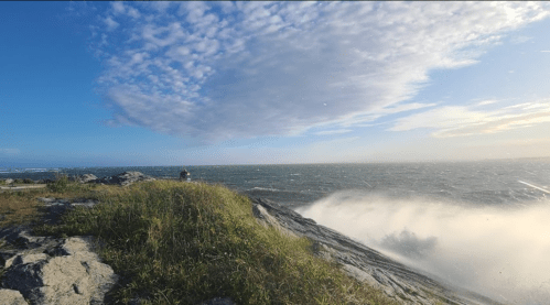 A person stands on rocky shore, overlooking turbulent waves under a blue sky with scattered clouds.