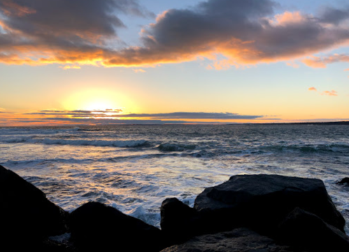Sunset over the ocean, with waves crashing against dark rocks and colorful clouds in the sky.