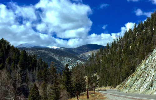 Scenic mountain landscape with lush trees, a winding road, and a blue sky filled with fluffy clouds.