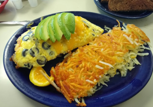 A blue plate with a cheese and olive omelet topped with avocado slices, alongside crispy hash browns and a lemon wedge.