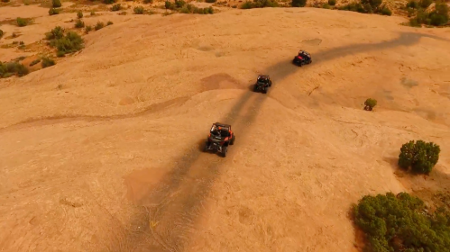 Four off-road vehicles driving on a sandy terrain under a clear sky, casting long shadows on the ground.