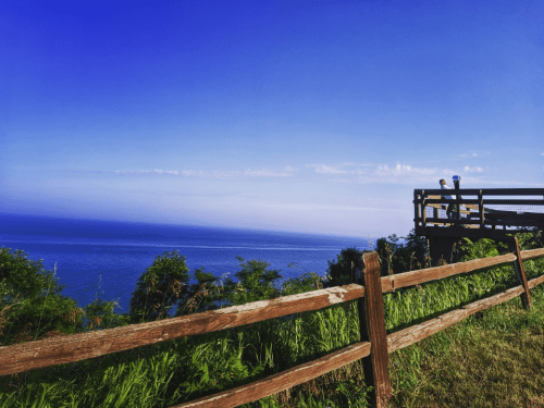 A scenic view of the ocean from a grassy hillside, with a wooden fence in the foreground and a clear blue sky.