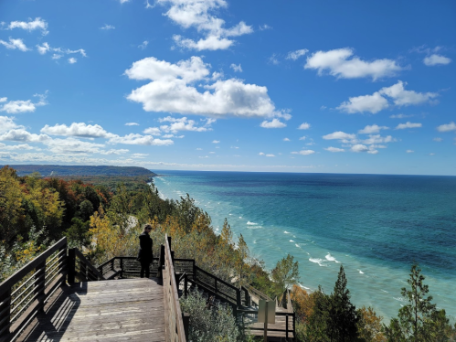 A person stands on a wooden overlook, gazing at a vibrant blue lake and colorful autumn trees under a partly cloudy sky.