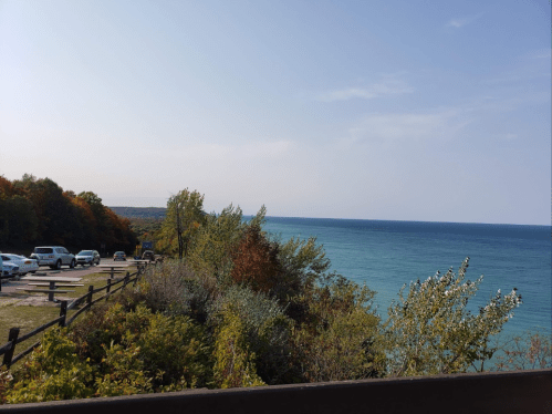 A scenic view of a calm blue lake surrounded by trees and a parking area on a sunny day.