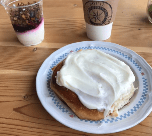A plate with a frosted cinnamon roll, a cup of yogurt with granola, and a coffee cup on a wooden table.