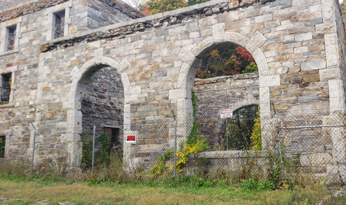 Abandoned stone building with arched windows, surrounded by overgrown grass and a chain-link fence.