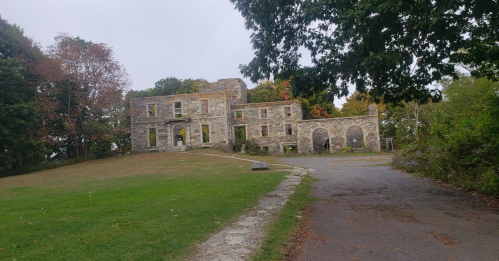 A stone building with multiple windows, surrounded by trees and grass, on a cloudy day.