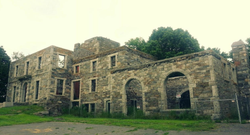 Ruins of a stone building with arched windows, surrounded by overgrown grass and trees under a cloudy sky.