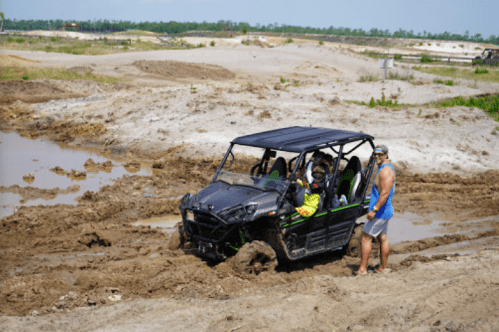 A man assists a group in a black off-road vehicle stuck in muddy terrain near a water-filled area.