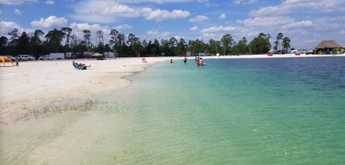 A serene beach scene with clear turquoise water, white sand, and people enjoying the sun and water under a blue sky.