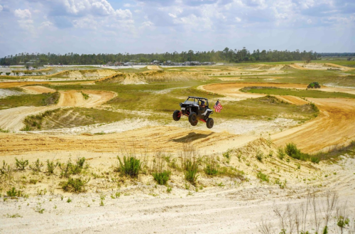A vehicle jumps over a dirt track, with a flag waving, surrounded by grassy hills and a clear blue sky.