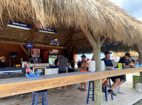 A beach bar with thatched roof, patrons enjoying drinks, and a lively atmosphere by the water.