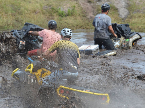 Two riders on ATVs splash through muddy water, creating a spray of mud in a vibrant outdoor setting.