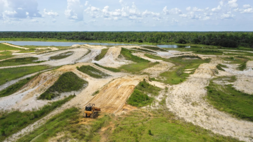 Aerial view of a sandy landscape with machinery, surrounded by greenery and a body of water in the background.