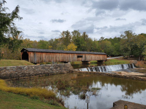 A wooden covered bridge spans a river with a small waterfall, surrounded by trees and cloudy skies.
