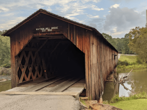 A wooden covered bridge with a sign reading "Watson Mill Bridge, Est. 1885," spanning a calm river.