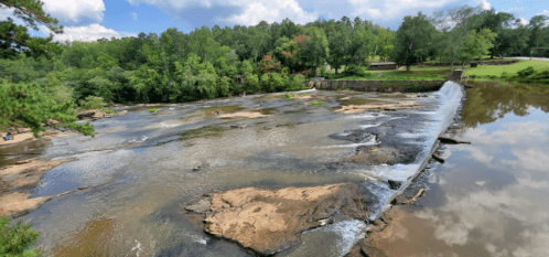 A serene river scene with a small waterfall, rocky banks, and lush greenery under a partly cloudy sky.
