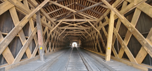 Interior view of a wooden covered bridge, showcasing a crisscross pattern of beams and a wooden floor leading to light.