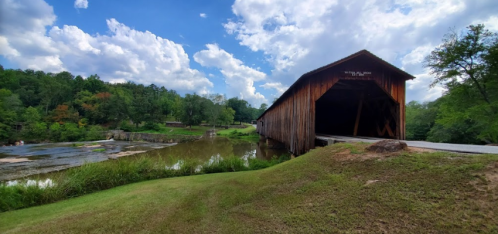 A wooden covered bridge beside a calm river, surrounded by lush greenery and a blue sky with fluffy clouds.
