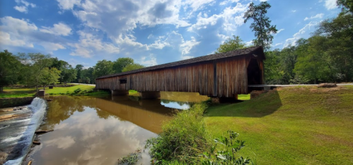 A wooden covered bridge spans a calm river, surrounded by lush greenery and a bright blue sky with scattered clouds.