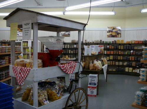 A quaint store interior featuring a wooden cart with a red grinder, surrounded by shelves of various snacks and treats.
