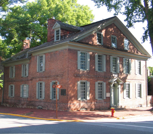 Historic brick house with green shutters, large windows, and a sloped roof, surrounded by trees and a sidewalk.