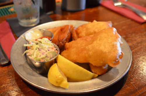 A metal plate with fried fish, coleslaw, sweet potato fries, and lemon wedges.