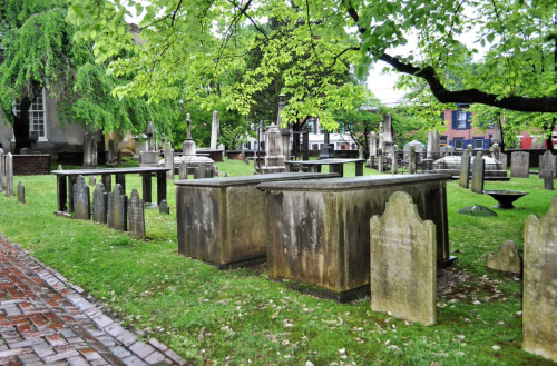 A peaceful cemetery with stone tombs and gravestones surrounded by lush green trees and a brick pathway.