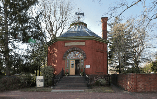A red brick building with a circular design, a green roof, and a chimney, surrounded by trees and a pathway.