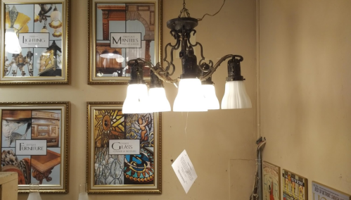 A vintage chandelier with four white glass shades hangs against a wall of framed art and decor samples.