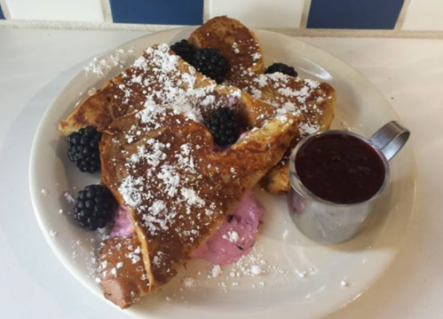 A plate of French toast topped with powdered sugar, blackberries, and a small cup of berry sauce.