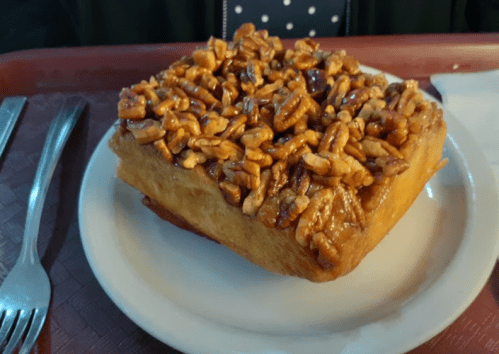 A square pastry topped with caramelized pecans, served on a white plate with a fork and knife nearby.