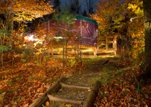A cozy yurt surrounded by autumn foliage, with a path leading through colorful fallen leaves at dusk.
