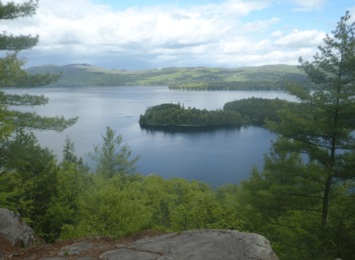 A serene lake view with an island surrounded by lush greenery and distant hills under a partly cloudy sky.