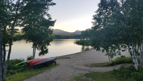 A serene lakeside scene with kayaks on the shore, surrounded by trees and mountains in the background at sunset.