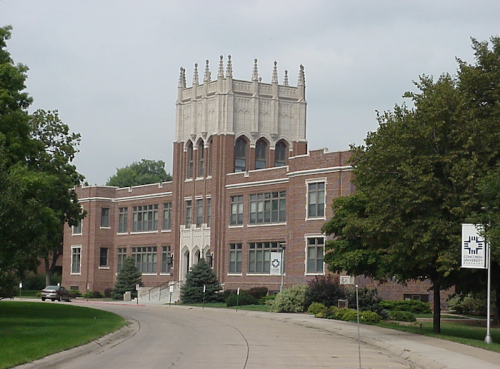 Historic brick building with a tall, ornate tower, surrounded by trees and a winding road. Overcast sky above.