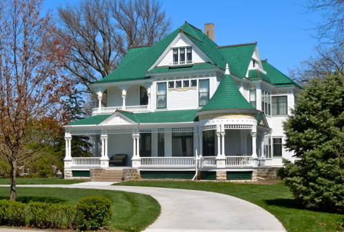A large Victorian-style house with a green roof, white trim, and a spacious porch, surrounded by manicured lawns.