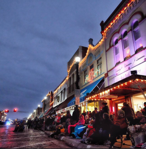 A festive street scene at dusk, with people gathered on the sidewalk, decorated buildings, and holiday lights.