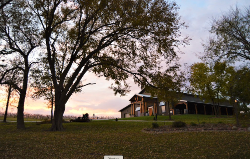 A large tree in the foreground with a rustic building and colorful sunset in the background.